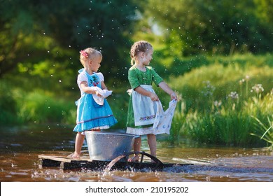 Two Cute Little Girls Doing The Laundry In A Small Aluminium Basin Standing On A Pontoon At The Pond In A Sunny Summer Day. Kids Are Playing.