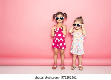 Two cute little girls standing in jumpers and sun glasses on the pink background in the studio. Summer, fun, family and vacations concept. Two fashion sisters posing. 