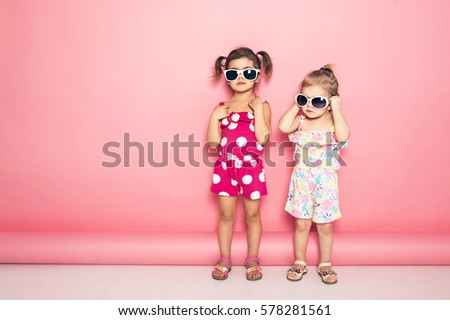 Two cute little girls standing in jumpers and sun glasses on the pink background in the studio. Summer, fun, family and vacations concept. Two fashion sisters posing. 