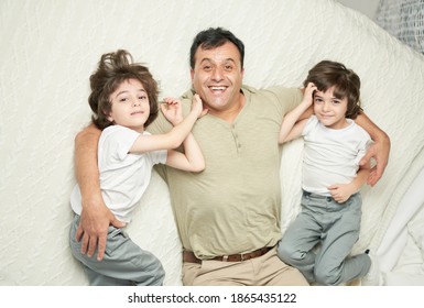 Two cute little latin boys, children and their father smiling at camera while lying together on a bed at home