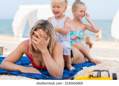 Two cute little sibling girls enjoy having fun playing sitting on tired exhausted mother's back at sea ocean beach. Frustrated mom make face palm gesture. Vacation family small kids trouble concept