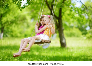 Two cute little sisters having fun on a swing together in beautiful summer garden on warm and sunny day outdoors. Active summer leisure for kids.