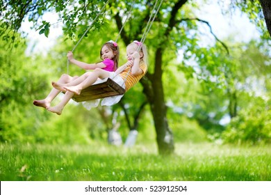 Two Cute Little Sisters Having Fun On A Swing Together In Beautiful Summer Garden On Warm And Sunny Day Outdoors 