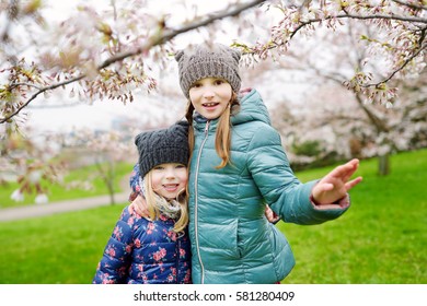 Two Cute Little Sisters Having Fun In Blooming Cherry Garden On Beautiful Early Spring Day 