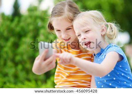 Two cute little sisters playing outdoor mobile game on their smart phones. Kids catching virtual pocket monsters. Modern addictive multiplayer location-based games.