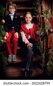 Two Cute Modern Kids In Elegant Classic School Uniform And Glasses Are Sitting On The Stairs In A Luxurious Vintage Interior. Christmas Decoration. Kid's School Fashion.