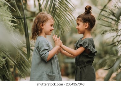 Two cute smiling little girls belonging to different races, in linen clothes, holding hands and walking in the botanical garden. Children explore tropical plants and flowers in the greenhouse. - Powered by Shutterstock