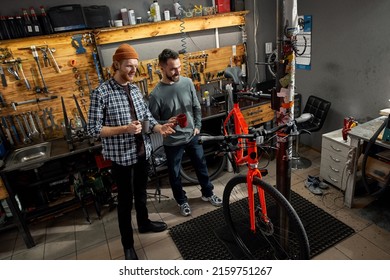 Two cycling mechanics checking bicycle and talking in workshop. Young caucasian men with cups of coffee or tea. Teamwork. Bike service, repair and upgrade. Garage interior with tools and equipment - Powered by Shutterstock