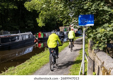 Two cyclist in fluorescent jackets and helmets riding on the tow path of a canal with their shadows on the ground. Narrow boats moored on the opposite bank of the canal, with reflections on the water.