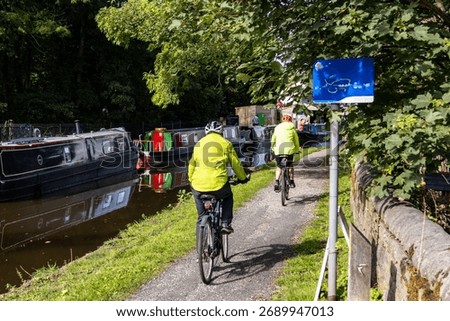 Two cyclist in fluorescent jackets and helmets riding on the tow path of a canal with their shadows on the ground. Narrow boats moored on the opposite bank of the canal, with reflections on the water.