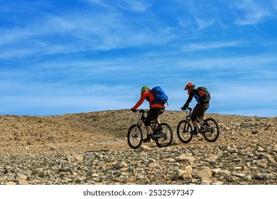 Two cyclists riding on rocky terrain under a bright blue sky, wearing helmets and backpacks. Mountain biking adventure. - Powered by Shutterstock