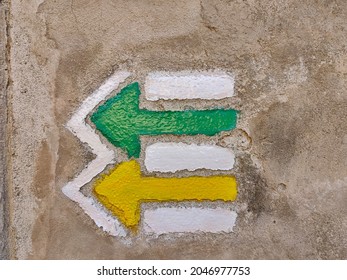 Two Czech tourist directional arrows painted on gray wall. One is green and the other is yellow. They both go in the same direction.