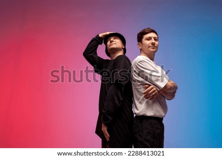 two dancer guys stand together in neon club lighting, male rap performers stand on red blue background and look up