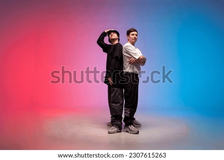 two dancer guys stand together in neon club lighting, male rap performers stand on red blue background and look up