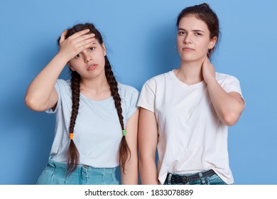 Two Dark Haired Sisters Posing Indoors Against Blue Wall, Sad Girl With Health Problems, Stressed Female Colleague Has Pain In Neck And Headache,look At Camera With Upset And Tired Look.