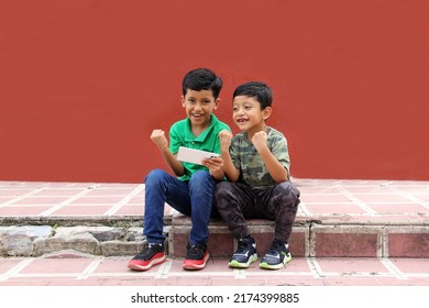 Two Dark-haired Latino Children Who Are Brothers Or Friends Who Live In Poverty Play Video Games With A Cell Phone, Make Video Calls And Take Selfies
