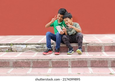 Two Dark-haired Latino Children Who Are Brothers Or Friends Who Live In Poverty Play Video Games With A Cell Phone, Make Video Calls And Take Selfies
