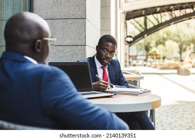 Two dark-skinned African American businessmen in suits and glasses with briefcases sit at a table in an outdoor city cafe. - Powered by Shutterstock