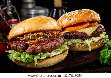 Two delicious beef burgers with salad trimmings, one a cheeseburger and the other with ham and ketchup on a dark wooden board in a close up view