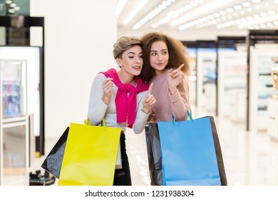 Two Delighted Diverse Young Women Being In Great Mood While Doing Shopping In A Big City Mall. Young Blonde Woman Showing Something In The Shop Showcase To Her Female Friend With Brown Frizzy Hair