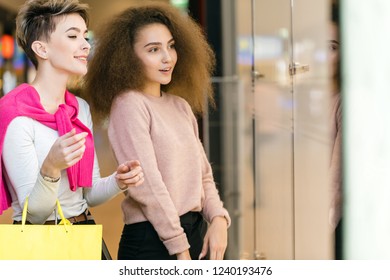 Two Delighted Diverse Young Women Being In Great Mood While Doing Shopping In A Big City Mall. Young Blonde Woman Showing Something In The Shop Showcase To Her Female Friend With Brown Frizzy Hair