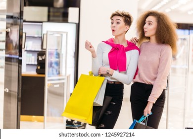 Two Delighted Diverse Young Women Being In Great Mood While Doing Shopping In A Big City Mall. Young Blonde Woman Showing Something In The Shop Showcase To Her Female Friend With Brown Frizzy Hair