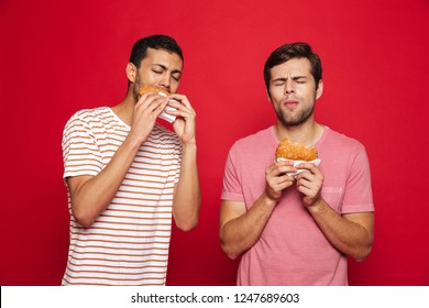 Two Delighted Men Friends Standing Isolated Over Red Background, Eating Burgers