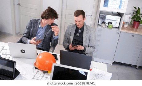 Two design engineers are sitting at a table with laptops in the meeting room and looking at their mobile phones. Young professionals. Break during the briefing at the architectural bureau.