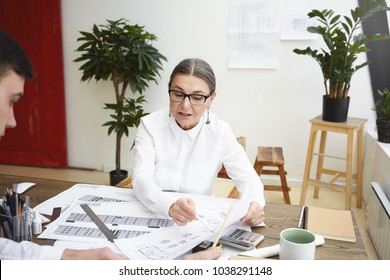 Two Designers Working On Blueprints Of Construction Project In Office. Senior Female Architect In Glasses Checking Drawings Of Her Young Male Assistant, Making Corrections, Sharing Ac Ideas And Vision