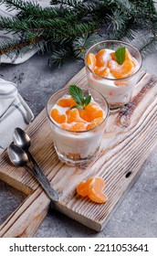 Two desserts in glass cups with cream and tangerines on a wooden board. On the background are branches of a green Christmas tree. - Shutterstock ID 2211053641