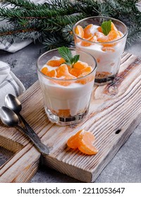 Two desserts in glass cups with cream and tangerines on a wooden board. On the background are branches of a green Christmas tree. - Shutterstock ID 2211053647