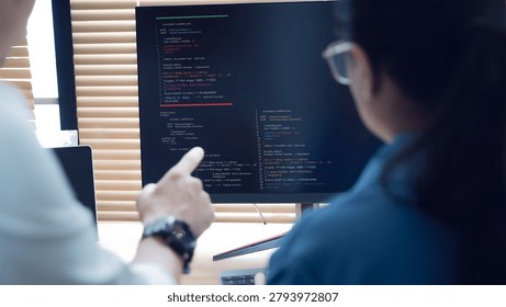 Two developers collaborate at a desk. One points at code on a large screen. Their focus shows teamwork and problem-solving. Soft lighting enhances the focused atmosphere. Ideal for tech, development
