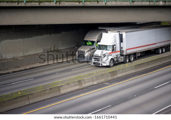 Two different industrial grade big rig semi trucks with
diesel engines and refrigerator semi trailers transporting
commercial cargo running on the divided highway under the concrete
bridge 