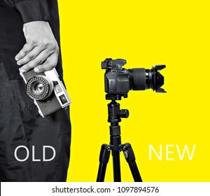 Two Different Photo Cameras, A Man Hand Holding Old Vintage Camera And New Camera With Tripod On Yellow Background. Comparison Of Different Generations Of Photographing Equipment. Technology Concept.