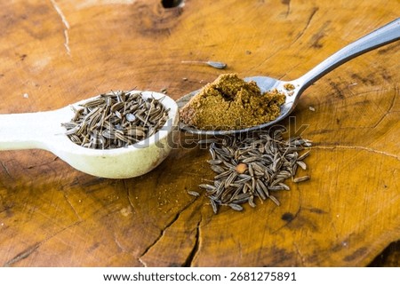Two different types of teaspoons, one filled with cumin seeds and one filled with ground cumin powder on a wooden background.