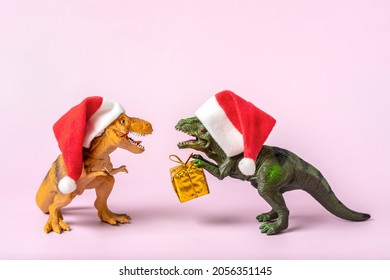 Two dinosaurs Rex in red Santa Claus hat holds golden gift box in its paws on pink background New Years Eve or Christmas Eve Art holiday card Creative idea for Merry xmas concept
