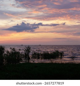 Two distant boats float on a serene lake under a vivid purple and orange sunset, with grass and reeds in foreground.