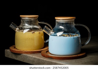 Two distinct herbal concoctions sit elegantly in stylish teapots, one a warm golden hue and the other a serene blue.