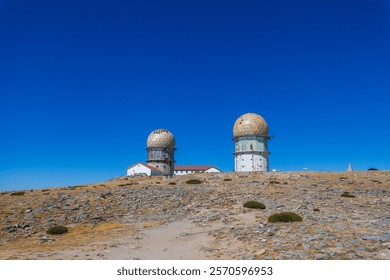 Two distinctive domed structures stand atop a barren, rocky hill against a clear blue sky, creating a striking architectural and natural contrast