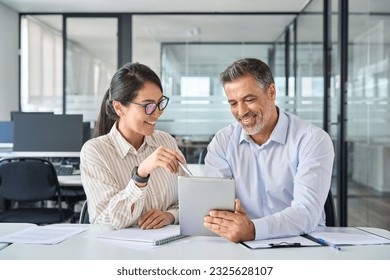 Two diverse business team people talking and working in office on corporate project at meeting. Young Asian employee and mid aged Latin executive looking at digital tablet company technology device. - Powered by Shutterstock