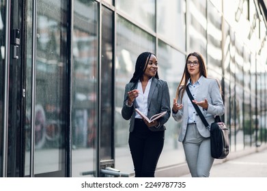Two diverse business woman meeting outside of the office. African american female project manager in motion outside the office building and discussing work with her caucasian female coworker. - Shutterstock ID 2249787735