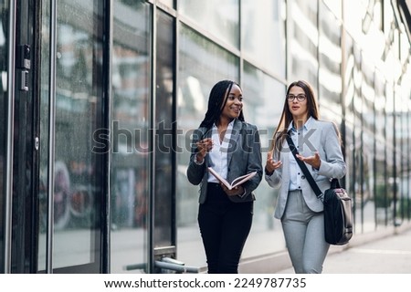 Two diverse business woman meeting outside of the office. African american female project manager in motion outside the office building and discussing work with her caucasian female coworker.