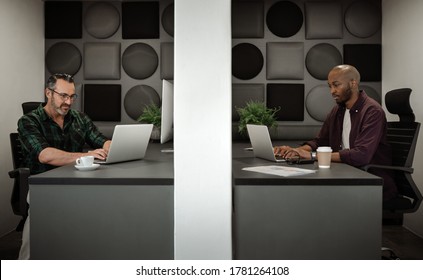 Two Diverse Businessman Working On Laptops In Separate Cubicles While Physical Distancing In The Workplace