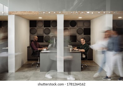 Two Diverse Businessmen Working At Desks In Cubicles With Blurred Colleagues Walking Around A Busy Office