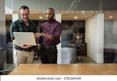 Two diverse businessmen working on a laptop together while standing at a desk in a busy modern office - Powered by Shutterstock