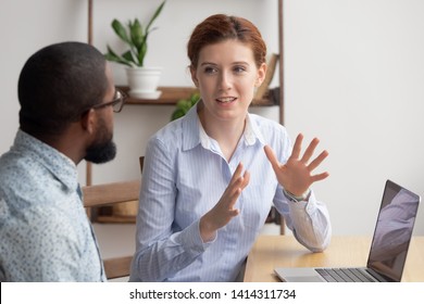 Two diverse businesspeople chatting sitting behind laptop in office. Excited caucasian female sharing ideas or startup business plan with black male coworker. Informal conversation, work break concept