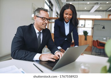 Two Diverse Businesspeople Smiling And Talking Together While Working On A Laptop At A Table In An Office