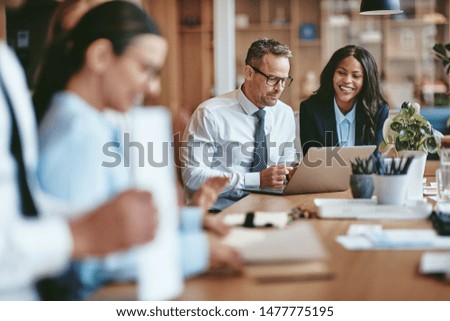 Two diverse businesspeople smiling while working on a laptop together at the end of a boardroom table in an office