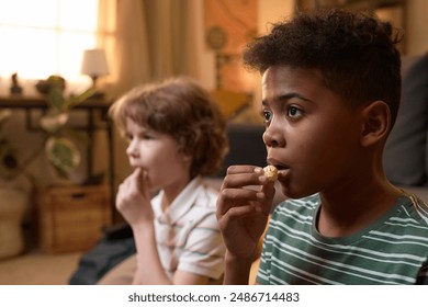 Two diverse children sitting on floor eating popcorn while watching TV in cozy old-fashioned living room with focused facial expression - Powered by Shutterstock