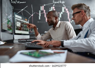 Two Diverse Colleagues Traders Talking To Each Other, Looking At Graphs While Sitting In The Office In Front Of Multiple Computer Screens. Stock Trading, People, Business Concept.